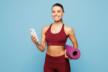 Young fitness trainer instructor sporty woman sportsman wears red sportswear training in home gym hold yoga mat use mobile cell phone isolated on plain blue background. Workout sport fit abs concept.
