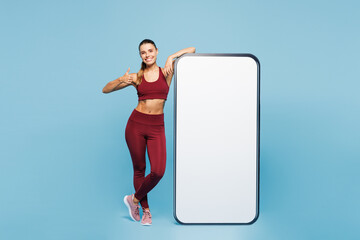 Full body young fitness trainer sporty woman in red sportswear train in home gym big huge blank screen mobile cell phone show thumb up isolated on plain blue background Workout sport fit abs concept