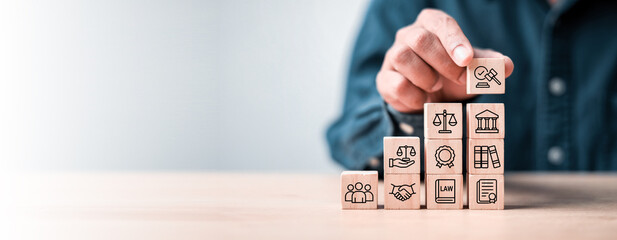 Business hand stacking wooden blocks with law and justice icons on light background. Concept of legal service, justice system, compliance, and business law consulting.