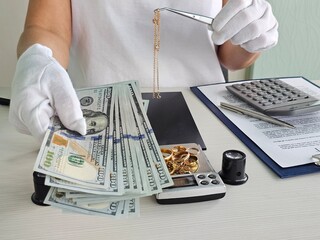 A person wearing gloves counts stacks of cash while examining gold jewelry on a balance scale in a retail setting. A calculator and documents are nearby, indicating financial activity concept
