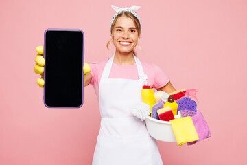 Young woman wears white apron hold basin with detergent bottles while doing housework tidy up use blank screen mobile cell phone isolated on plain pastel light pink background. Housekeeping concept.