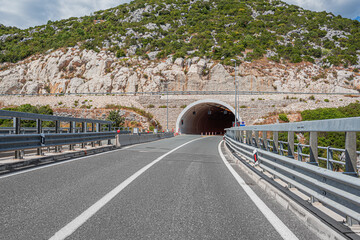 A two-lane road with guardrails on both sides leads into a tunnel carved through a large, rocky hill covered in green vegetation under a cloudy sky.