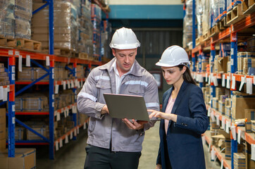 Business woman in suit and safety helmet review inventory record on laptop computer. Stand in a well-organized warehouse. Shelve are lined with various product, indicating a busy working environment