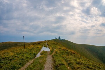 Hiking trail leading to a mountain summit with a radar or communication station under dramatic clouds. Remote scenic landscape in the hills after rain.