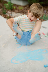 A young boy plays on a pavement, painting with bright chalk using his feet. He laughs joyfully as he explores his artistic side, surrounded by colorful designs and a vibrant outdoor setting.