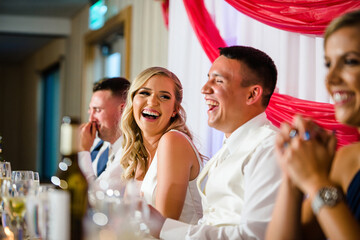 A joyful blonde bride and a groom laughing heartily at their wedding reception, surrounded by happy guests. A candid moment celebrating love and happiness.