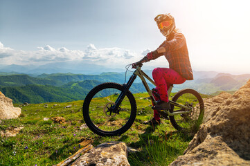 Naklejka premium Tourist with bicycle enjoying valley view from mountain top. Male cyclist riding mountain bike outdoors. Male tourist standing on grassy path in mountains wearing helmet