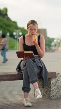Woman seated outdoors with crossed legs, holding a book in one hand while eating snacks and reading, the blurred background features a lamp post lined up with a blur view of a woman walking