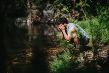 A man crouches by a forest stream, washing his face with water. The scene is peaceful and serene,...