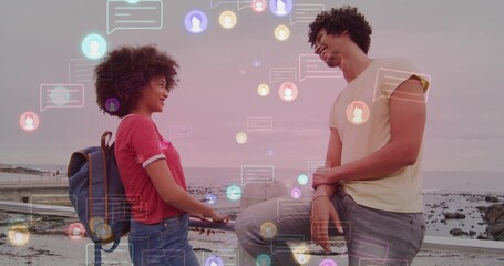 Exchanging messages, couple standing on seaside walkway with metal railing, backpack and chat icons