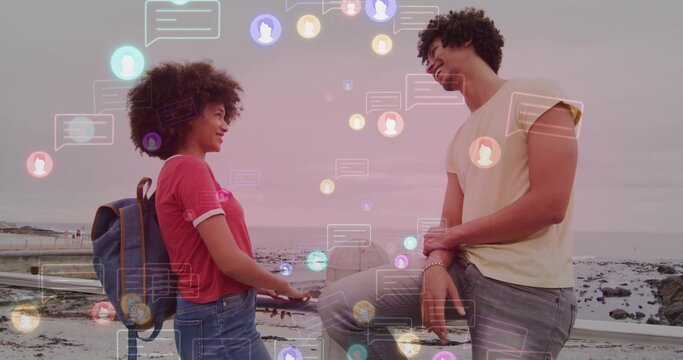 Exchanging messages, couple standing on seaside walkway with metal railing, backpack and chat icons