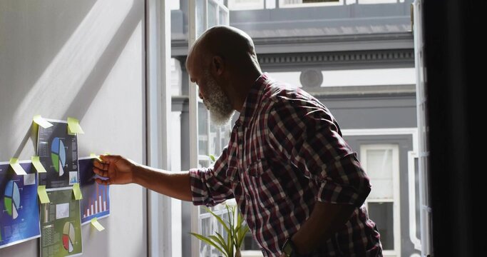 Senior man attaching printed charts to gray wall in office beside window, with yellow sticky notes - Powered by Adobe