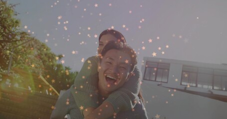 Laughing Korean man giving piggyback ride to woman in modern white house backyard, with lens flares