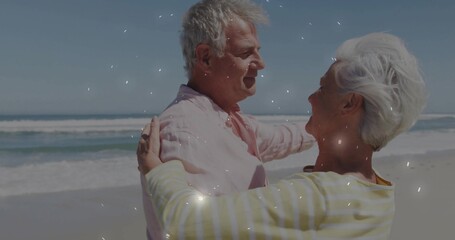 Embracing senior couple standing on sandy beach, smiling in pastel pink shirt and striped top