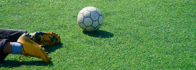 Worn soccer ball on green artificial turf beside a player’s yellow cleats, pause after training;...