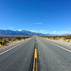 Straight road leads the eye to distant mountains under a vast blue sky. Desert landscape fringes the road. Peaceful travel