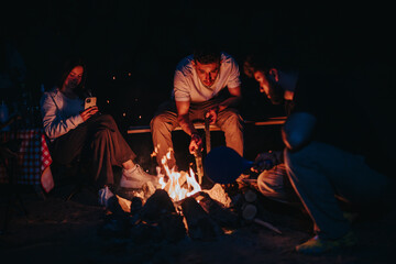 A group of friends gathering around a campfire during the night, enjoying warmth and companionship,...