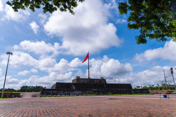 A red and yellow star flag flies proudly above a historic citadel in Hue, Vietnam, framed by lush green trees and a bright, cloudy sky.