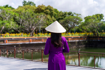 A woman in a vibrant purple ao dai and conical hat gazes over a tranquil moat in Hue, with sunflowers and green trees in the peaceful background.