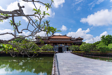 A paved stone walkway crosses a serene moat towards a magnificent Hue Imperial City palace, framed by tree branches and set against a bright, cloudy sky.