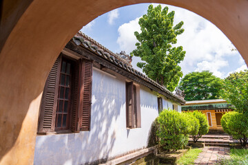 A close-up view through an arched entryway reveals a traditional building with wooden shutters and a classic tiled roof, surrounded by lush garden greenery under a bright sky.