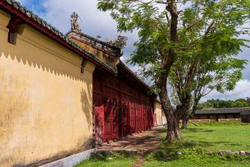 A vibrant yellow wall with striking red wooden doors and a traditional roofline stands beside a lush green lawn and trees, capturing a classic Hue scene.