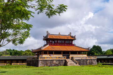 A classic two-story pavilion with an orange roof and stone base stands on a green lawn in Hue, framed by tree branches under a bright, cloudy sky.