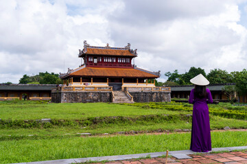 A woman in a purple ao dai and conical hat admires an ancient Hue royal palace, a beautiful blend of traditional architecture and lush greenery under a cloudy sky.