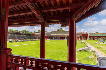 A peaceful courtyard in the Hue Imperial City, featuring a large green lawn, traditional buildings, and weathered stone walls, is viewed from under a red-pillared pavilion.