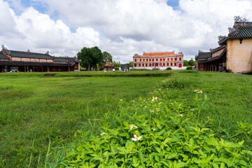 A colorful and ornate imperial palace with bright red windows stands behind a green field dotted with white wildflowers, under a cloudy sky in Hue.