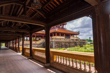 A tranquil garden courtyard with traditional buildings and manicured hedges is viewed through the dark wooden corridor of an ancient palace in Hue under a cloudy sky.