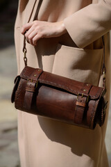 A professional fashion shot capturing a female model showcasing a cylindrical brown leather bag with detailed buckles and a strap, highlighting her beige coat.