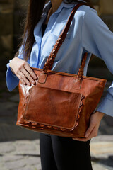 A professional close-up shot of a model showcasing a refined brown leather tote bag with unique braided handles and a decorative front pocket