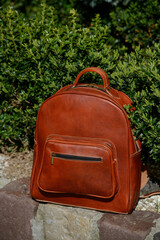 A high-quality brown leather backpack with a front zipper pocket is showcased on a stone ledge in a natural, outdoor setting, highlighting its rugged elegance.