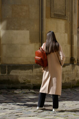 A beautiful fair-skinned model with long, dark hair is photographed from the back, showcasing a sophisticated brown leather backpack and a timeless beige trench coat.