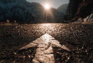 Low-angle shot looking down a textured road toward snowy mountains with a bright sunburst peaking over the horizon, focused road with blurred bg