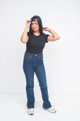 An edgy young Asian woman in a full body shot standing with a confident smirk while fixing her cap worn backward with attitude. Isolated on a white background.