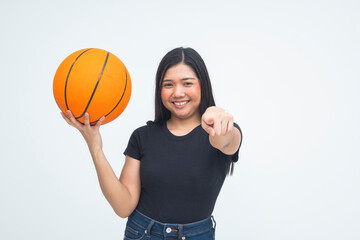 A bold young Asian woman in a half body shot smiling with pride while holding a basketball in one hand and pointing directly at the camera challengingly. Isolated on a white background.