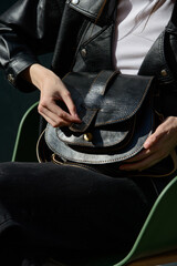 A stylish model in a black leather jacket showcases a unique black leather cross-body bag with a glossy sheen and a front flap pocket