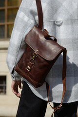 A stylish model is photographed from the back, showcasing a classic brown leather backpack and a trendy plaid overshirt against an urban backdrop