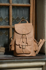 A high-quality beige leather backpack with detailed buckles and pockets is presented on a rustic windowsill, highlighting its elegant, vintage design.