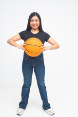 A joyful young Asian woman in a full body shot standing confidently while holding a basketball with both hands in front of her and smiling. Isolated on a white background.