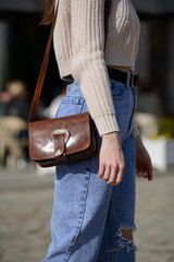 A stylish model in a knit sweater and ripped jeans showcases a chic brown leather cross-body bag...