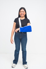 An optimistic young Asian woman in a full body shot standing with a bright smile while wearing an arm sling despite her injury. Isolated on a white background.