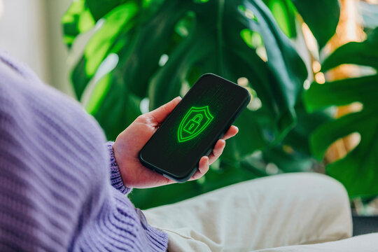 A young woman is holding a smartphone in her hands, and on the screen a glowing cybersecurity lock symbol is displayed - Powered by Adobe