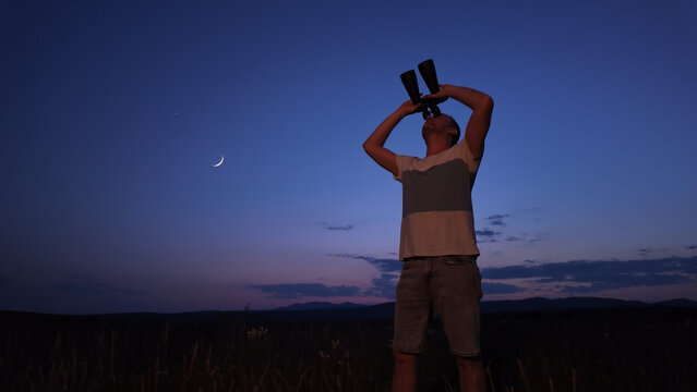 Amateur astronomer using binoculars for watching stars, Moon, planets and other celestial objects from a countryside field in nature.