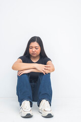 A troubled young Asian woman in a half body shot sitting on the floor, hugging her knees and gazing down with sadness and unhappiness. Isolated on a white background.