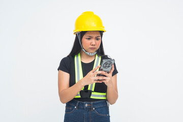 An irritated young Asian woman in a half body shot holding her phone while wearing a protective helmet, displaying a mad and displeased look. Isolated on a white background.
