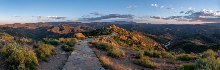 Panoramic landscape showing a stone path ascending a hill with rolling mountains under a cloud-flecked sky during a golden sunset