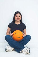 A bubbly young Asian woman in a half body shot sitting on the floor with legs crossed, smiling and holding a basketball with a focused and joyful demeanor. Isolated on a white background.
